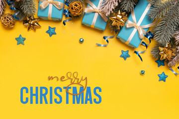 Wall Mural - Merry Christmas text on yellow background with pine tree, blue gifts and festive decorations top view. Xmas greeting card