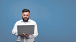 © artdolgov - A bearded man holds a laptop in his hands on a blue background. A solid man in a white shirt works at a laptop and looks into the camera.
