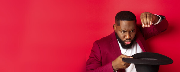 Wall Mural - Close-up of charismatic Black male magician perform a trick with his hat, performing on christmas party, standing over red background