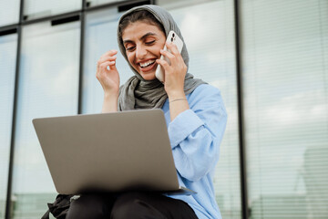 Cheerful muslim woman talking on cellphone and using laptop while sitting outdoors