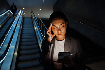 Wall Mural - Young asian woman frowning while using smartphone standing on escalator indoors