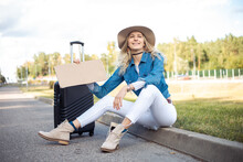Smiling Solitude Blond Woman In Hat Hitchhiking, Sit On Curb Near Grass, Hold Blank Carton Board With Suitcase Baggage