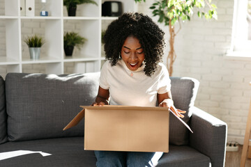 Young african woman sit on couch at home unpacking parcel cardboard box with online purchase