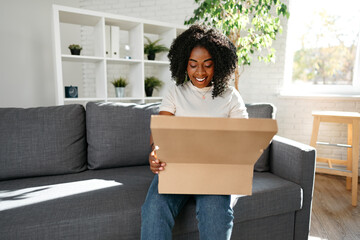 Young african woman sit on couch at home unpacking parcel cardboard box with online purchase