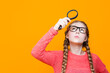 Calm Intelligent Caucasian Girl in Glasses And Coral Knitted Sweater Holding Big Magnifying Glass While Searching And Looking Upwards On Yellow.