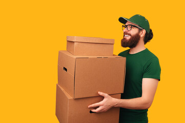 Strong bearded man is holding up some boxes and looking at unseen client over yellow background.