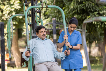 A physiotherapist or professional caregiver watches closely as a senior adult woman exercises on an outdoor gym.