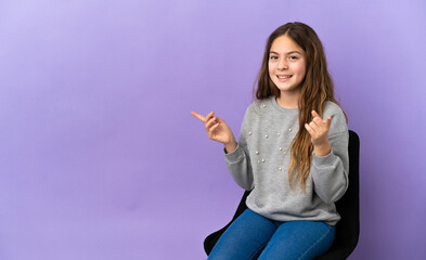 Canvas Print - Little caucasian girl sitting on a chair isolated on purple background pointing finger to the laterals and happy