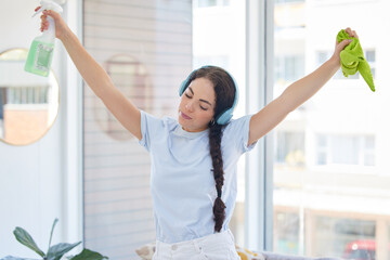 Poster - Woman cleaning, dancing and listening to music with headphones while holding cloth and detergent to clean with energy, fun and audio podcast. Female cleaner or maid working in a house or apartment