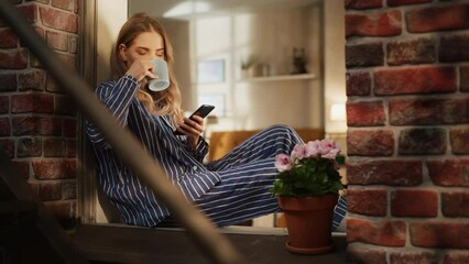 Poster - Young Beautiful Woman Using Smartphone and Enjoying her Morning Coffee While Sitting on her Bedroom Windowsill. Caucasian Female in Striped Pyjamas Doing her Online Shopping and Relaxing