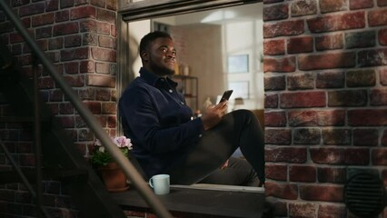 Poster - Black Male in Comfortable Casual Clothes Using Smartphone and Sitting on his Windowsill. Handsome Young Man Browsing Internet and Smiling While Watching Funny Videos and Commenting on a Website