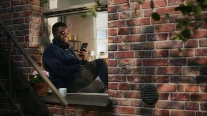 Poster - African American Young Man in Comfortable Casual Clothes Using Smartphone and Sitting on his Windowsill. Black Male Staying Connected and Using Internet in a Brooklyn Style Brownstone House