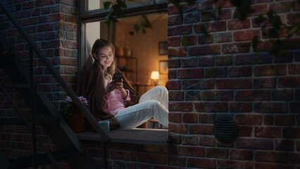 Aufkleber - Happy Beautiful White Woman Using her Smartphone While Sitting on her Windowsill During Evening. She's Browsing the Internet and Checking Videos on Social Networks and Having Fun