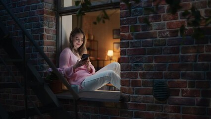 Wall Mural - Beautiful White Woman Using Smartphone While Sitting on an Open Windowsill. Attractive Girl with Healthy Creative Lifestyle Relaxes at Home in the Evening, Spending Some Alone Self-care Time