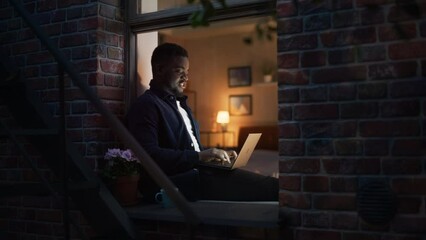 Poster - Handsome, Smiling, Black African Man Works on a Laptop While Sitting at Windowsill at Home. Portrait of a Young Male Freelancer Working on a Computer and Enjoying his Time, Making Progress on Project