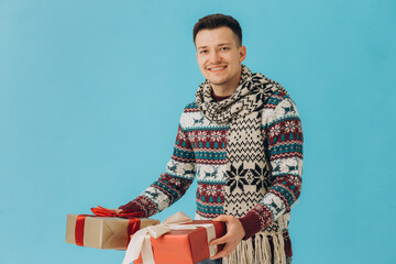 Young man in Christmas sweater and scarf holding many gift boxes with gift ribbon bow isolated on blue background. Happy new year, celebration concept.