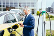 © Iryna - Mature cauvasian businesss man using smart phone and waiting power supply connect to electric vehicles for charging the battery in car outdoor.