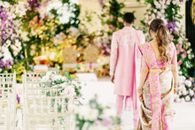 Back View Of Groom And Bride At The Decorated Hall At Wedding Ceremony. Elegant Couple Of Newlyweds Dressed In Light Pink Ceremonial Costumes, Standing In Hall Decorated With Fresh Flowers