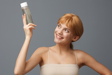 pretty woman in a white T-shirt with light clean skin, with red silky hair gathered in a ponytail on a gray background with a transparent bottle of water.Horizontal studio shot.