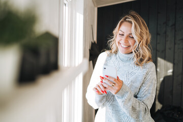 Young woman with blonde curly hair in grey sweater using mobile phone in hands near window at house