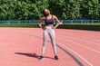 © DimaBerlin - African American woman wearing grey leggings and black top prepares for competition. Black sportswoman stretches body with hands on hips for running marathon on red jogging track in sunny morning