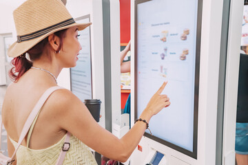 A female customer uses a touchscreen terminal or self-service kiosk to order at a fast food restaurant. Automated machine and electronic payment