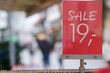 Nahaufnahme eines roten Preisschildes mit dem Aufdruck „Sale“ und der Zahl „19“ auf einem Kleiderständer vor einem Geschäft mit Menschen in der Fußgängerzone im verschwommenen Hintergrund