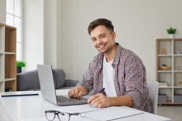 Wall Mural - Portrait of happy male student working on his computer. Handsome young man looks at camera and smiles while sitting at table with laptop and writing in notepad. Studying online concept