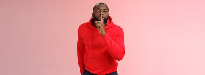 Wall Mural - Shhh it taboo. Portrait sensual funny cute african-american bearded man in red hoodie bending camera show shush gesture index finger on lips telling secret asking keep quiet not tell anyone mystery