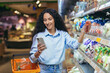 © Liubomir - A Latin American woman is standing in a supermarket with a basket near the refrigerator with vegetables. Holds the phone in his hands, reads the list, composition of products, chooses cherry tomatoes.