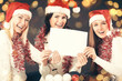 © yurolaitsalbert - three young girls in costume of Santa Claus showing blank sheets. Beautiful bokeh effect from a garland.