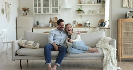 Wall Mural - Cheerful millennial couple use new mobile application resting on cozy sofa with smartphone, staring at cellphone screen make self-portraits having fun on internet on leisure at home. Modern tech usage