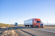 Team of two orange and blue big rigs semi trucks transporting cargo in dry van semi trailers running together on the flat straight highway road in California.
