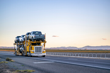 Yellow industrial car hauler big rig semi truck transporting pick up trucks on the two level semi trailer driving on the evening straight highway road