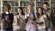 © fizkes - Happy multiethnic first year college students posing in university library, looking at camera, standing at bookshelves, showing like thumb up gestures, smiling, laughing, recommending education