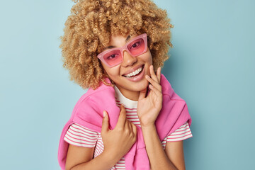 Poster - Indoor shot of pretty curly haired woman smiles gently touches face has healthy skin wears trendy sunglasses and caasual outfit isolated over blue background. Positive human emotions concept