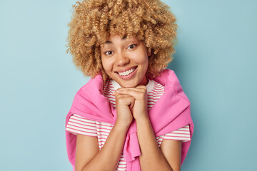 Wall Mural - Portrait of cheerful curly haired woman keeps hands under chin smiles gently shows white teeth dressed in casual striped t shirt and sweater tied over shoulders isolated over blue background
