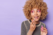 © wayhome.studio  - Photo of good looking young woman with curly hair applies lip gloss wears bright makeup undergoes beauty procedures before dating isolated over purple background blank space for your promotion