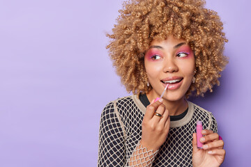 Wall Mural - Photo of good looking young woman with curly hair applies lip gloss wears bright makeup undergoes beauty procedures before dating isolated over purple background blank space for your promotion