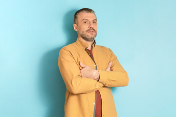 Poster - People emotions concept. Portrait of serious mature man posing with folded arms, standing on blue studio background