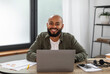 © Prostock-studio - Portrait of happy latin male freelancer sitting at table, working on laptop at home office interior, copy space