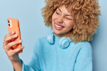 Wall Mural - Horizontal shot of cheerful friendly woman winks eye and takes selfie via smartphone enjoys pleasant distant conversation wears casual blue jumper and stereo headphones around neck stands indoor