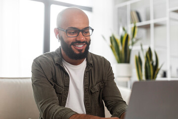 Wall Mural - Happy latin businessman working on laptop computer, wearing earbuds and looking at screen, sitting at home interior