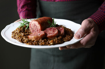 Cibo italiano. Mani uomo che tengono piatto bianco con cotechino servito con lenticchie. Piatto tipico di Capodanno. Avvicinamento.