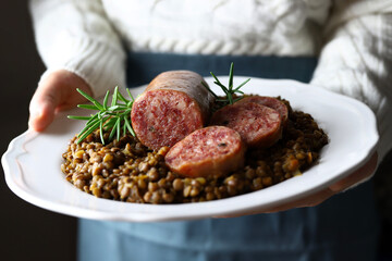 Cibo italiano. Mani donna che tengono piatto bianco con cotechino servito con lenticchie. Piatto tipico di Capodanno. Avvicinamento. 