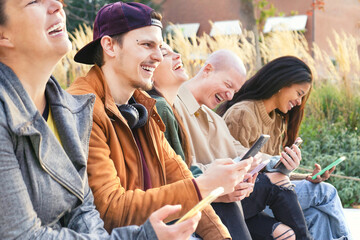 Young urban friends using smartphone outdoors sitting at park bench . Happy group of people laughing and sharing social content with mobile smart phone  at university college- Tech life style Concept