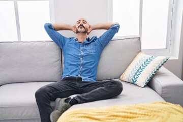 Wall Mural - Young hispanic man relaxed with hands on head sitting on sofa at home