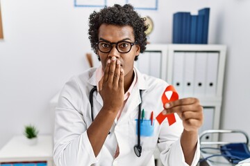 Poster - African doctor man holding support red ribbon covering mouth with hand, shocked and afraid for mistake. surprised expression