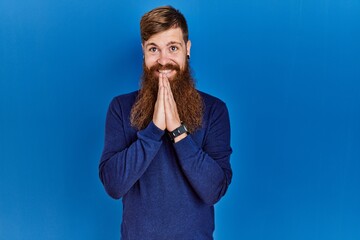 Canvas Print - Redhead man with long beard wearing casual blue sweater over blue background praying with hands together asking for forgiveness smiling confident.