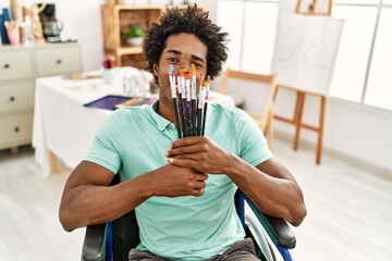 Poster - Young african american disabled artist man holding paintbrushes sitting on wheelchair at art studio.